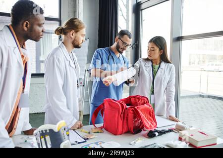 Gesundheitspersonal in Brille und Uniform auftragen Kompressionsstrumpf auf Arm einer asiatischen Frau in der Nähe von multiethnischen Studenten, erste-Hilfe-Kasten und medizinisch Stockfoto