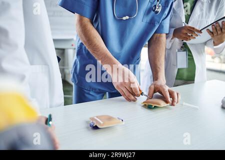 Erste-Hilfe-praxisorientiertes Lernen, zugeschnittene Ansicht des medizinischen Lehrers in Uniform Praktizieren mit Injektions-Trainingspad in der Nähe von Schülern in weißen Kitteln, Leben-s Stockfoto