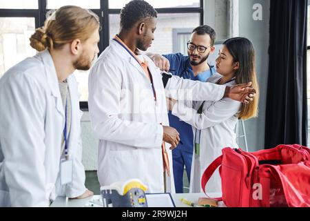 Gesundheitsfachkraft hilft asiatischen Studenten, die Kompressionsstrumpf auf den Arm eines afroamerikanischen Mannes in der Nähe von erste-Hilfe-Kasten und medizinische Ausrüstung auftragen, Leben- Stockfoto