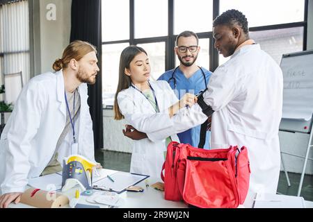 Junge asiatische Frau, die Druckstaub auf dem Arm eines afroamerikanischen Mannes in der Nähe des erste-Hilfe-Kastens, medizinische Ausrüstung und Arzt mit interrassistischen Stu anlegt Stockfoto