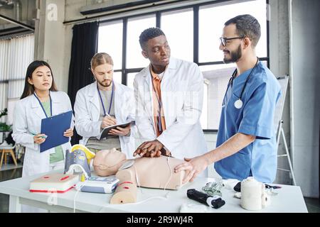 Positiver medizinischer Lehrer schaut auf afroamerikanischen Mann, der Thoraxkompressionen auf der HLW-Prüfpuppe in der Nähe von medizinischen Geräten und multiethnische Studen praktiziert Stockfoto
