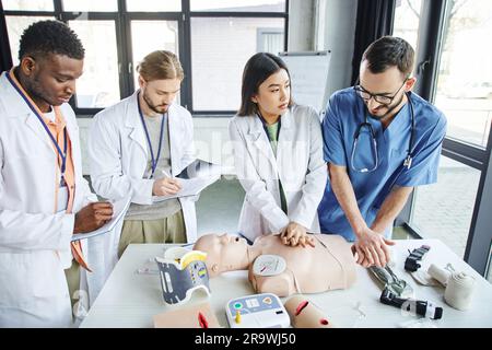 Multiethnische Studenten in weißen Kitteln schreiben in Notizbüchern in der Nähe von medizinischem Lehrer assistierende asiatische Frau, die Thoraxkompressionen an der HLW-Prüfpuppe macht, kommen Stockfoto