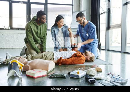 Medizinischer Lehrer, der Kompressionsturniquet auf Arm des Seminarteilnehmers in der Nähe einer asiatischen Frau, eines afroamerikanischen Mannes und medizinischer Ausrüstung im Zug anlegt Stockfoto