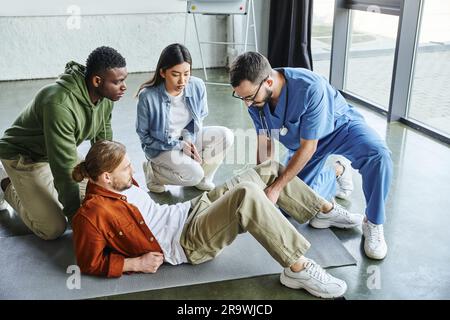 afroamerikanischer Mann und asiatische Frau, die einen medizinischen Lehrer anschauen, der Kompressionsverband auf das Bein des Teilnehmers des erste-Hilfe-Trainings Seminars anlegt, b Stockfoto