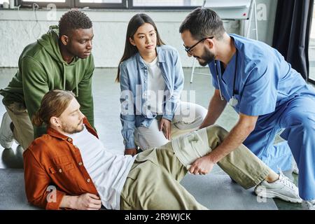 Pflegekraft, die während des erste-Hilfe-Seminars im Trainingsraum Kompressionsbandage am Bein des Mannes in der Nähe von interrassistischen Teilnehmern anlegt, Blutung verhindern Stockfoto