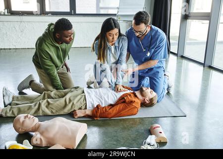 Junge asiatische Frau, die Thoraxkompressionen bei einem Mann in der Nähe von medizinischem Lehrer, HLW-Übungspuppe und afroamerikanischer Teilnehmer am erste-Hilfe-Training Seminar, Stockfoto