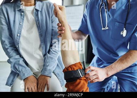 Schnittbild eines professionellen Sanitäters mit Kompressionsstrumpf am Arm eines Mannes in der Nähe einer jungen Frau während eines erste-Hilfe-Seminars im Trainingsraum Emerg Stockfoto