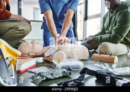 Partielle Ansicht eines Sanitäters, der Thoraxkompressionen an der HLW-Übungspuppe in der Nähe multiethnischer Teilnehmer und medizinischer Geräte, Defibrillator, Wundbehandlungssimull durchführt Stockfoto