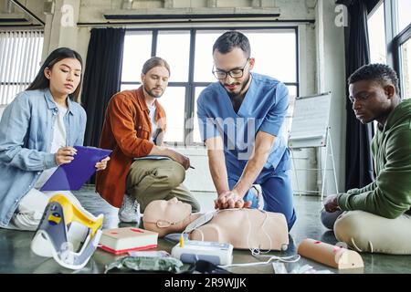 Junge asiatische Frau mit Klemmbrett, die einen Sanitäter ansieht, der Thoraxkompressionen auf der HLW-Prüfpuppe in der Nähe von medizinischen Geräten und multiethnische Teilnehmer von f durchführt Stockfoto