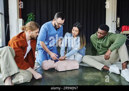 Junge asiatische Frau, die lebensrettende Fähigkeiten praktiziert, indem sie Thoraxkompressionen auf der HLW-Übungspuppe in der Nähe des multiethnischen Teams und medizinischen Lehrers, Kardiopulmone Stockfoto