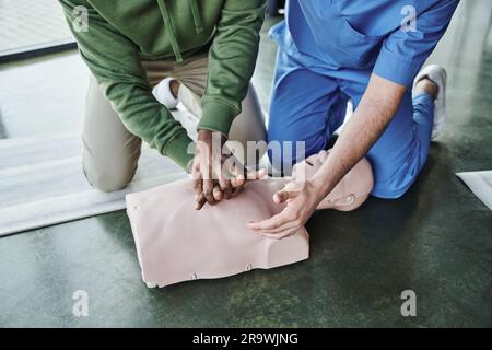 Zugeschnittener Blick auf einen afroamerikanischen Mann, der lebensrettende Fähigkeiten praktiziert und Thoraxkompressionen an der HLW-Prüfpuppe in der Nähe eines professionellen Sanitäters auf der ersten ai durchführt Stockfoto