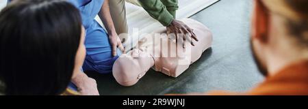 Teilansicht eines afroamerikanischen Mannes, der Thoraxkompressionen auf der HLW-Prüfpuppe in der Nähe des medizinischen Lehrers und Teilnehmer auf verschwommenem Vordergrund, Life-savi Stockfoto