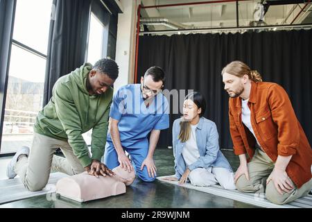 ein afroamerikanischer Mann führt Thoraxkompressionen an der HLW-Prüfpuppe durch, während er beim ersten-Hilfe-Seminar in der Nähe eines professionellen Sanitäters und einer vielfältigen Gro lernt Stockfoto