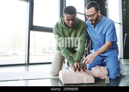 Medizinischer Lehrer in Uniform und Brille zeigt auf die HLW-Puppe während afroamerikanischer Mann Thoraxkompressionen, effektive lebensrettende Fähigkeiten Stockfoto
