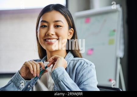 Porträt einer jungen und charmanten asiatischen Geschäftsfrau mit braunem Haar und einem fröhlichen Lächeln, die einen Stift hält und in einem modernen Büro in die Kamera schaut Stockfoto