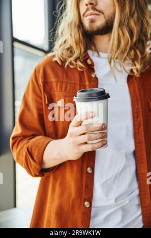 Kurzer Blick auf einen bärtigen Geschäftsmann mit langen Haaren, der einen Pappbecher mit einem Getränk zum Mitnehmen hielt, während er in einem modernen Büro stand, Hipster-Stil, Erfolg Stockfoto