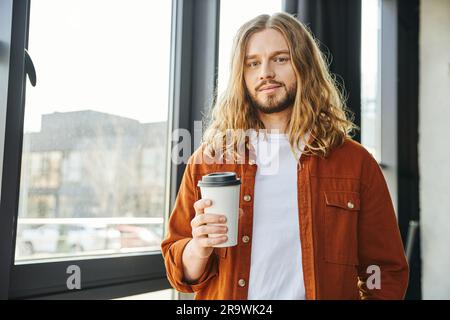 Attraktiver und cooler Geschäftsmann, der Pappbecher mit Kaffee zum Mitnehmen hielt, während er neben dem Fenster stand und in die Kamera schaute, in der Nähe des Fensters im Büro, lange Zeit Stockfoto