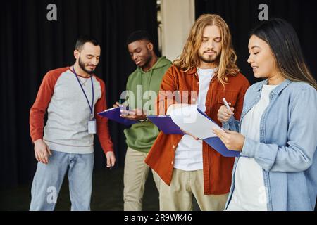 Medizinisches Seminar und erste-Hilfe-Training, junge asiatische Frau mit Klemmbrett, die mit einem langhaarigen Mann in der Nähe des Ausbilders und afroamerikanischer Teilnehmer spricht, Stockfoto