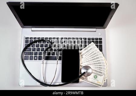 Mobiltelefon mit Dollarrechnungen und Stethoskop auf Laptop-Tastatur. Stethoskop auf Dollarscheinen auf Laptop-Tastatur Stockfoto