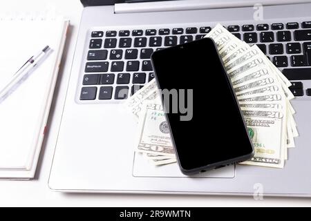 Mobiltelefon neben Dollarscheinen auf Laptop-Tastatur. Draufsicht auf Laptop mit Handy und Dollarrechnungen. Handy auf Laptop-Tastatur und Dollar Stockfoto