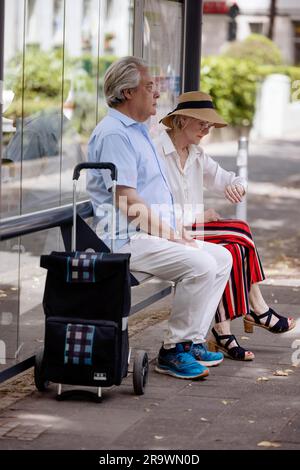 Ältere Ehepaare in Sommerkleidung mit einem Trolley, der an einer Bushaltestelle in Köln, Nordrhein-Westfalen, Deutschland, auf einen Bus wartet Stockfoto