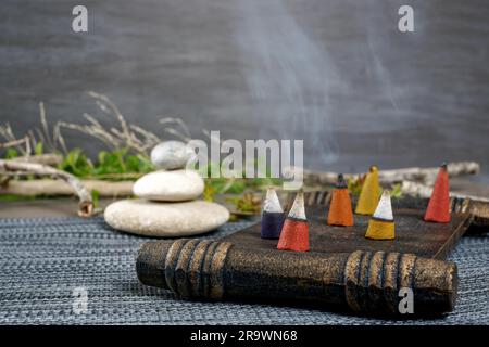 Farbenfrohe Räucherstäbchen brennen und rauchen auf einem Holztablett mit gestapelten Zen-Steinen Stockfoto