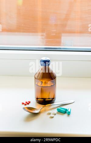 Eine Flasche Hustensaft mit einem Löffel und einige Pillen auf dem Tisch am Fenster Stockfoto