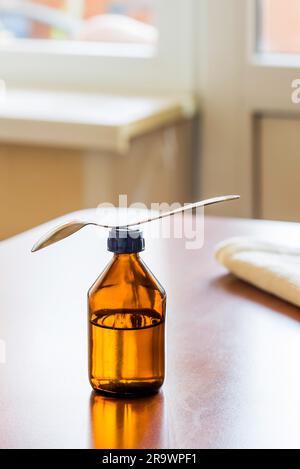 Eine Flasche Hustensaft mit einem Löffel auf dem Tisch am Fenster Stockfoto