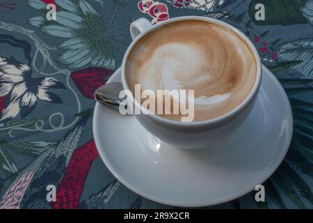 Blick von oben auf einen heißen Cappuccino, serviert in einer weißen Tasse auf einer Tischdecke mit einem von der Natur inspirierten Muster. Es ruft ein Gefühl hervor, wie man Ruß genießt Stockfoto