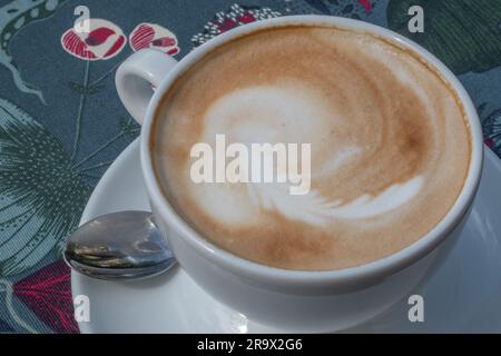 Blick von oben auf einen heißen Cappuccino, serviert in einer weißen Tasse auf einer Tischdecke mit einem von der Natur inspirierten Muster. Es ruft ein Gefühl hervor, wie man Ruß genießt Stockfoto