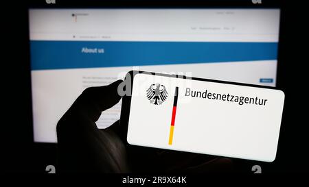 Person, die ein Mobiltelefon mit dem Logo der Bundesnetzagentur (BNetzA) auf dem Bildschirm vor der Webseite hält. Konzentrieren Sie sich auf das Display des Telefons. Stockfoto