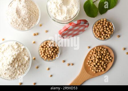 Details des ganzen und raffinierten Sojabohnenmehls auf einer weißen Küchenbank mit Glasschüsseln gefüllt mit Getreide und Mehl. Draufsicht. Stockfoto