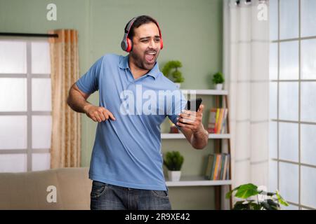 Fröhlicher, glücklicher indischer Mann, der tanzt, während er Musik über Kopfhörer hört, indem er sein Mobiltelefon zu Hause hält - Konzept des Vergnügens, des Wochenendlebens und Stockfoto