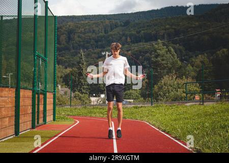 Ein braunhaariger Junge mit einer sportlichen Figur, die ein weißes T-Shirt und schwarze Shorts trägt, springt über ein athletisches Oval. Training zur Verbesserung des Springens, CO Stockfoto