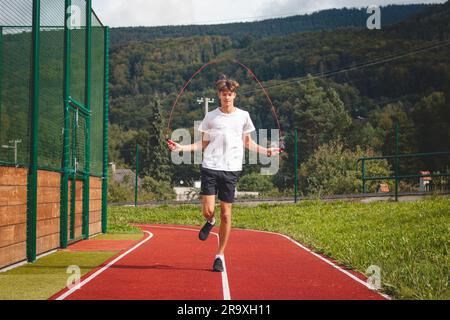 Ein braunhaariger Junge mit einer sportlichen Figur, die ein weißes T-Shirt und schwarze Shorts trägt, springt über ein athletisches Oval. Training zur Verbesserung des Springens, CO Stockfoto