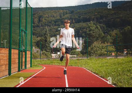 Ein braunhaariger Junge mit einer sportlichen Figur, die ein weißes T-Shirt und schwarze Shorts trägt, springt über ein athletisches Oval. Training zur Verbesserung des Springens, CO Stockfoto