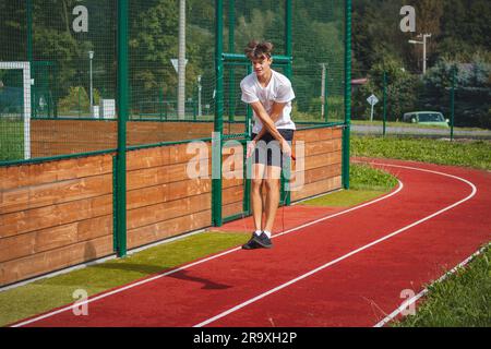 Ein braunhaariger Junge mit einer sportlichen Figur, die ein weißes T-Shirt und schwarze Shorts trägt, springt über ein athletisches Oval. Training zur Verbesserung des Springens, CO Stockfoto