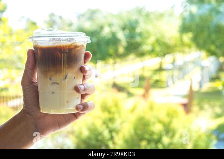 Eine Person hält einen erfrischenden Eiskaffee Latte mit verschiedenen Schichten Milch und Espresso in einem hellen, sonnigen Garten. Stockfoto