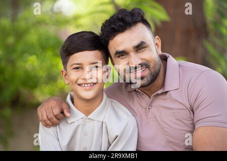 Glücklicher lächelnder indischer Vater mit Sohn, der sich gegenseitig umarmt, indem er vor die Kamera in den Park schaut - Konzept von Familienzusammengehörigkeit, Generation und Vaterschaft. Stockfoto