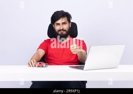 Junger Mann im roten Kleid, der auf dem Tisch sitzt, mit Auto und Laptop, der Daumen nach oben macht Stockfoto