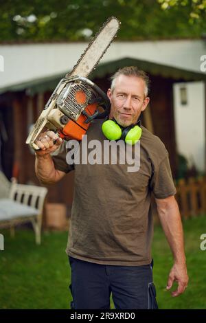 Gärtner mittleren Alters, der nach der Arbeit eine Kettensäge über die Schulter hält, mit Gehörschutz, der in die Kamera schaut Stockfoto