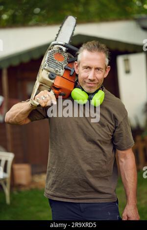 Gärtner mittleren Alters, der nach der Arbeit eine Kettensäge über die Schulter hält, mit Gehörschutz, der in die Kamera schaut Stockfoto