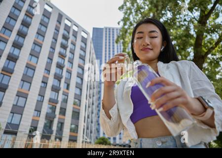 Fröhliche vietnamesische Frau, die im Freien Mineralwasser aus einer Glasflasche trinkt. Porträt einer schönen jungen südostasiatischen Frau, die hydriert Stockfoto