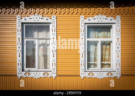 Zwei Fenster mit geschnitzten Holzjammen, die mit traditionellen weißen Mustern an der Fassade eines gelb gestrichenen russischen isba dekoriert sind Stockfoto