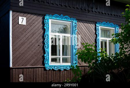 Zwei Fenster mit blau geschnitzten Holzjammen, die mit traditionellen Mustern an der Fassade eines russischen isba, einem aus Holz erbauten Haus in Russland, dekoriert sind Stockfoto