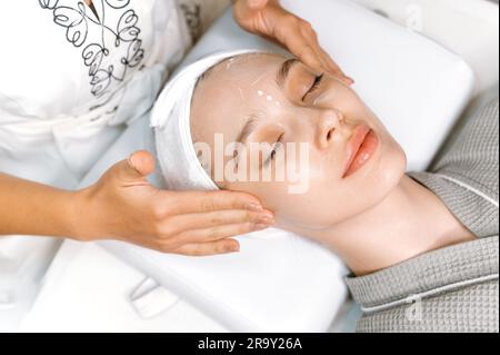 Gesichtspflege. Eine kaukasische, entspannte Frau mit geschlossenen Augen kümmert sich um die Behandlung im Salon, erhält eine Gesichtsmassage mit feuchtigkeitsspendender Creme, kümmert sich um die Jugend der Haut, beugt Falten vor Stockfoto