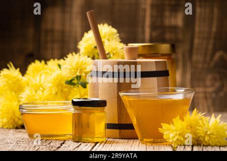 Kreative Zusammensetzung von Honig in einem Glasgefäß und einer Schüssel, einem Holzdipper und gelben Blumen auf einem alterlichen Holzhintergrund. Stockfoto