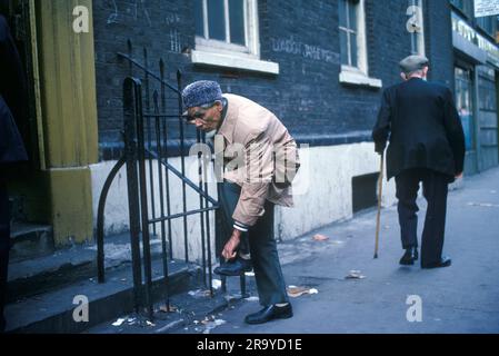 Whitechapel, Brick Lane, London 1970er, Vereinigtes Königreich. Ein alter muslimischer Mann zieht seine Schuhe aus, bevor er die Jamme-Masjid-Moschee in Brick Lane betritt, während ein alter weißer Mann mit Gehstock und flacher Mütze vorbeiläuft. Whitechapel, London, England, ca. 1978. 70s HOMER SYKES Stockfoto