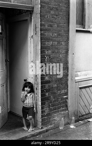 East End London 1970er, The Peabody Estate in Princess Street, ein multiethnischer Wohnblock in 1970er Großbritannien. Ein neues Immigrantenkind steht in der Tür zum Wohnblock. Zwei leere Milchflaschen aus Glas befinden sich draußen auf dem Bürgersteig. Whitechapel, London, England, ca. 1975. 70S GB HOMER SYKES Stockfoto