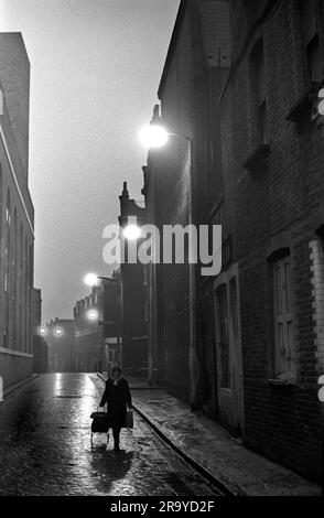 East End London 1970s, Brick Lane, eine Frau zieht ihre Einkaufstüte entlang der Kopfsteinpflasterstraße an viktorianischen Wohnblocks vorbei. Peabody Estate Tower Hamlets, London, England um 1976. HOMER SYKES AUS DEN 70ER JAHREN Stockfoto